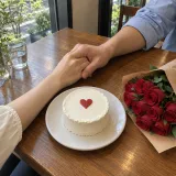 Clasped hands on a cafe table next to a small cake with a red heart and a bouquet of red roses