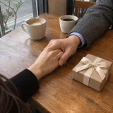 Two people holding hands at a cafe table beside two cups of coffee and a wrapped gift box