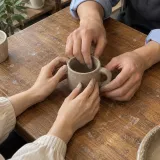 Two people shaping a clay mug together on a wooden table